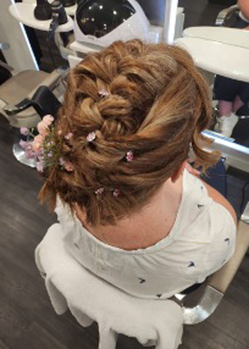 A woman is sat in a salon chair and has a plait updo with pink and purple flowers in her hair.
