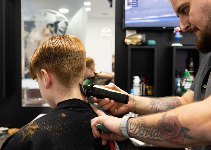 A boy is sat in a barbers chair and a barber is using clippers on his hair.