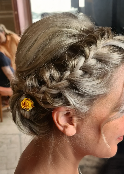 A bride with a plait going into a low bun hair style with a small yellow flower at the back.