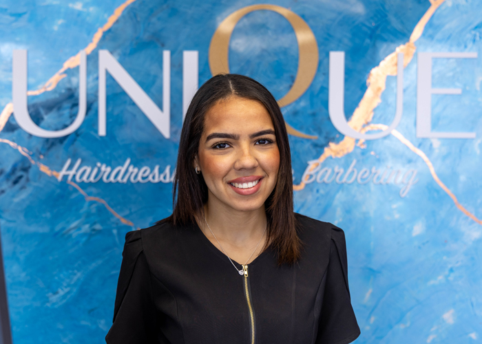 Sasha Kandiji from Unique Hairdressing & Barbering smiling in front of a blue wall.