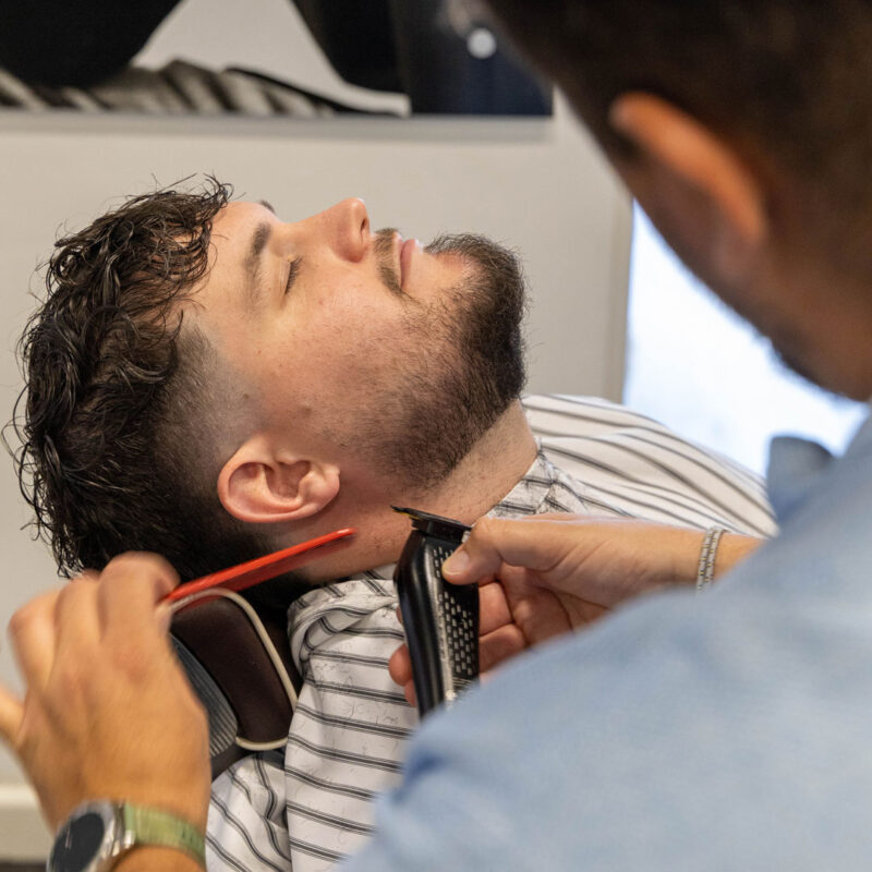 A man with his head laid back, getting his beard trimmed by a barber at Unique.