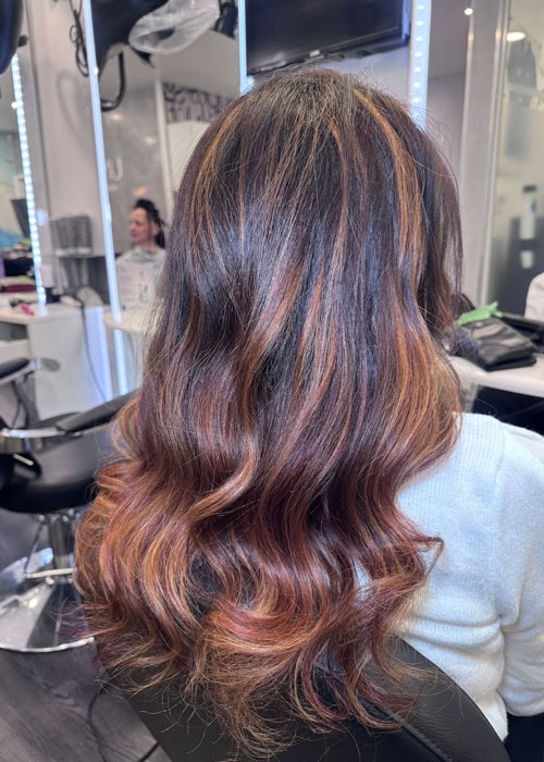A girl sat in a salon chair with dark hair and light red low lights.