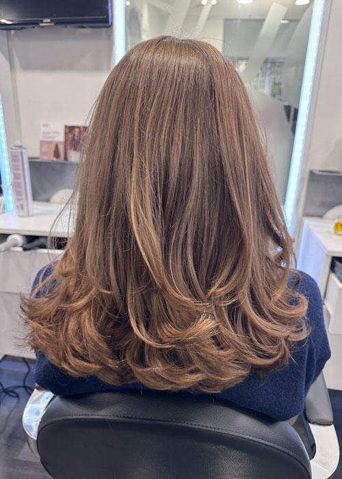 A girl sat in a hair salon chair with brunette layered hair that has been blow-dried.